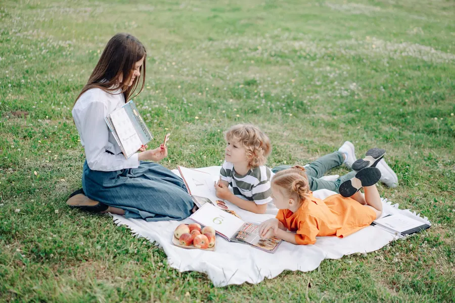 A woman sitting on a blanket in a grassy field, reading aloud from a book with two young children lying beside her.