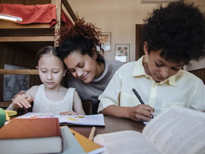 A mother leaning over a wooden table, helping two children as they write and work through books at home.